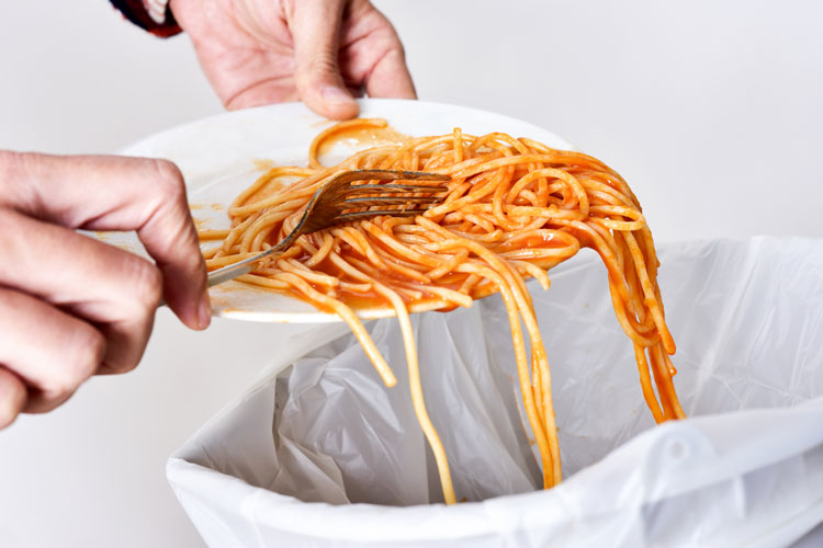 Ett par händer som skrapar ner spaghetti från en tallrik ner i en soptunna.
