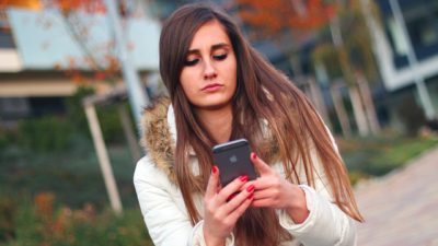 En kvinna med vit jacka och långt brunt hår håller i och tittar på sin smartphone.