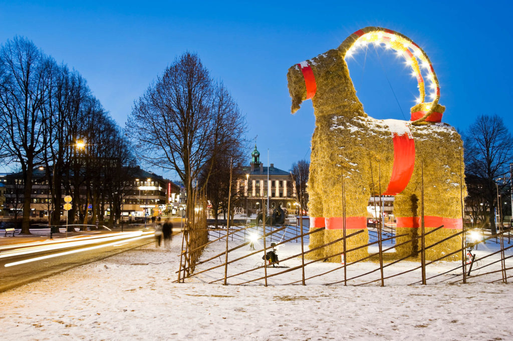 En stor bock av halm står bredvid flera träd. Bocken har röda band runt magen och benen. Runt bocken är det ett staket. På marken är det snö.