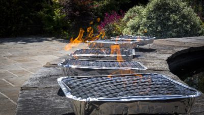 Bild på fem engångsgrillar som står uppradade på en stenmur. Det kommer lågor från grillarna.